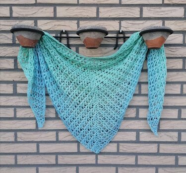 Gehäkeltes Dreieckstuch in Türkis‑ und Blautönen mit offenem Muster, an einer Ziegelwand hängend.