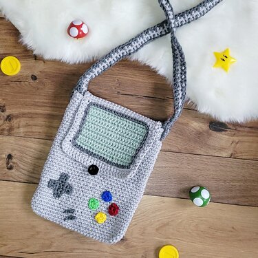Hellgraue gehäkelte Umhängetasche im Retro-Konsolen-Look mit mintgrünem Bildschirmfeld und bunten Tasten.