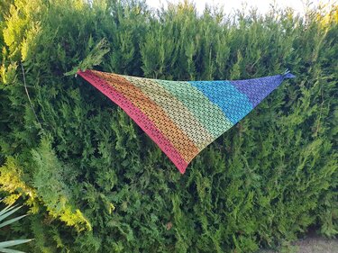 Gehäkeltes dreieckiges Tuch in Regenbogenfarben, aufgehängt an einer grünen Hecke