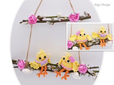 Handgehäkelte gelbe Küken mit lila Details auf einem Zweig, geschmückt mit Weidenkätzchen und rosa Stoffblüten vor hellem Hintergrund.