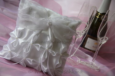 Weißes Ringkissen mit Faltornamenten, Perlenmitten und Schleifenbändern neben Gläsern