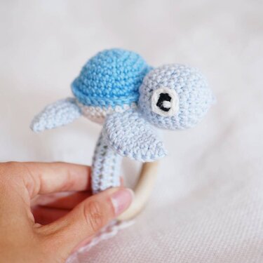 Hand holding pale blue crocheted turtle rattle attached to a wooden ring