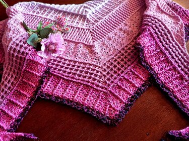 Nahaufnahme eines gehäkelten Dreieckstuchs in Pink- und Lilatönen auf Holz; unterschiedliche Häkelmuster sind gut erkennbar.