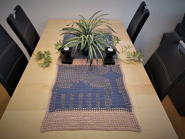Gehäkelter Tischläufer in Blaugrau und Beige mit stilisierter Tiersilhouette auf einem hellen Holztisch, dekoriert mit einer Grünpflanze und Kerzenhaltern.