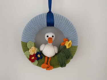 Gehäkelter Türkranz in Hellblau und Grün mit kleiner weißer Ente, bunten Häkelblüten, grünen Blättern und blauem Aufhängeband.