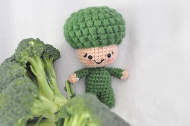 Gehäkeltes grünes Brokkoli-Amigurumi mit lächelndem Gesicht neben frischem Brokkoli