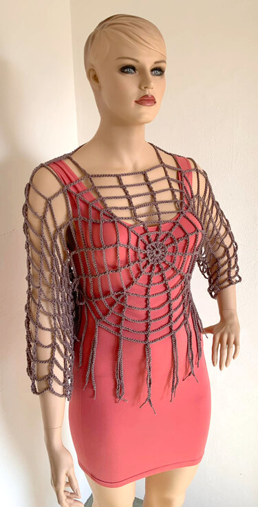 Schaufensterpuppe trägt graues gehäkeltes Spinnennetz-Oberteil mit Fransen über korallfarbenem Kleid.