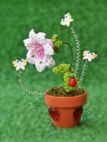 Gehäkelte weiße und rosa Glockenblume in einem Terrakotta-Topf mit roten Herzsteinen, Schmetterlingen auf Drahtspiralen und einem Marienkäfer vor grünem Hintergrund.
