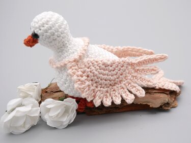 Gehäkelte weiße Taube mit zartrosa Flügeln auf Rindenstück neben weißen Rosen