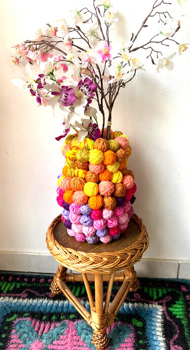 Bunte Vase aus Garnknäueln, gefüllt mit weißen und lila Kunstblumen, auf einem Rattanhocker vor weißer Wand.