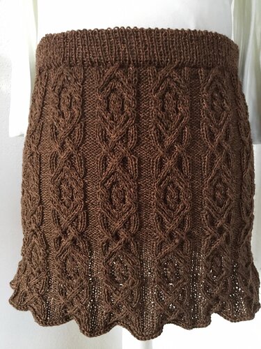 Dunkelbrauner Strickrock mit detailreichem Zopfmuster und gewelltem Saum vor hellem Hintergrund.