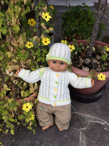 Puppe mit weißer Strickjacke und passender Mütze im Lochmuster, im Freien zwischen gelben Blüten