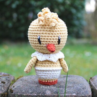 Gehäkeltes Amigurumi-Küken in Hellgelb mit weißem Kragen und orangefarbenem Schnabel, stehend auf einem Stein.