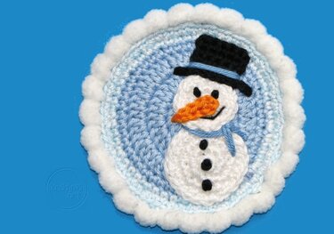 Runde gehäkelte Schneemann-Applikation mit schwarzem Zylinder, orangefarbener Nase und blauem Schal auf hellblauem Hintergrund mit weißem Bommelrand.