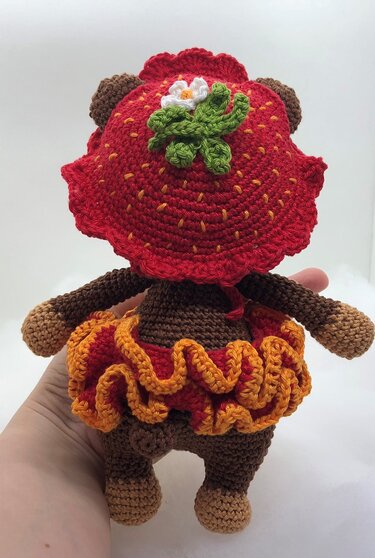 Rückansicht eines Amigurumi-Teddybären mit Erdbeer-Mütze und rot-orangefarbenem Rüschenrock