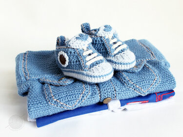 Zwei blaue gehäkelte Babysneaker mit weißen Schnürbändern auf gefalteter blauer Babyjacke und Bodysuit