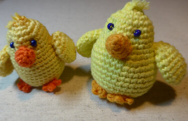 Zwei gelbe gehäkelte Küken-Amigurumi mit orangefarbenen Schnäbeln und blauen Perlenaugen auf hellem Untergrund.