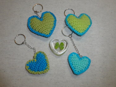 Vier gehäkelte Herz-Schlüsselanhänger in Blau- und Grüntönen mit Metallringen; mittig ein Glasherz mit einem vierblättrigen Kleeblatt.