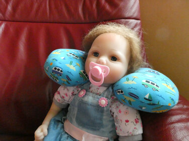 Puppe mit blauem U-förmigem Nackenkissen mit Wohnmobil- und Bootsmotiven auf rotem Sessel