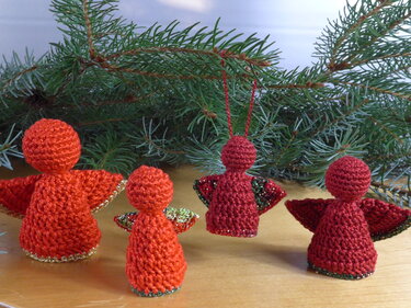 Vier gehäkelte rote Weihnachtsengel, drei stehend und einer hängend, vor Tannenzweigen auf einer Holzfläche.