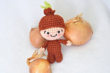 Gehäkelte Amigurumi-Puppe im braunen Zwiebelkostüm zwischen echten Zwiebeln
