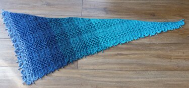 Gehäkelter dreieckiger Drachenschwanz-Schal mit blau-türkis Farbverlauf und Muschelkante auf Holzboden