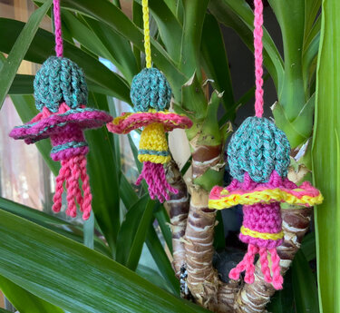 Drei gehäkelte, quallenartige Ornamente in Türkis, Pink und Gelb hängen an grünen Blättern einer Zimmerpflanze.