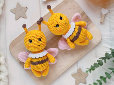 Two yellow and brown striped crocheted amigurumi bees with pink wings and white collars on a wooden board.