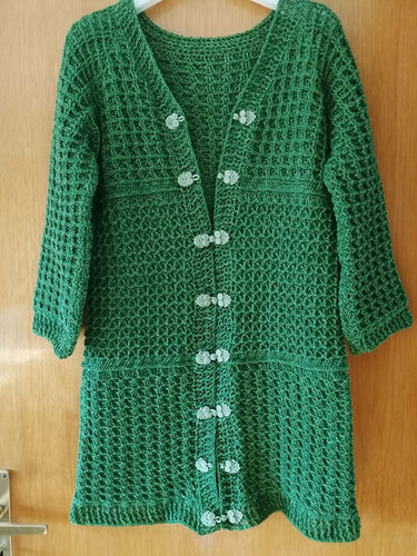 Grüne handgehäkelte lange Damenjacke mit V‑Ausschnitt und floralen Knöpfen, an einem Bügel vor Holztür hängend