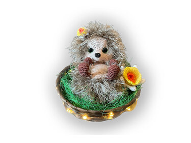 Gehäkelter Amigurumi-Igel mit Fransengarn-Stacheln im geflochtenen Körbchen, mit grünem Deko-Gras, Lichterkette und gelb-weißer Häkelblume.