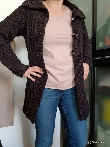 Lange dunkelbraune Häkeljacke mit breitem Kragen und Knebelverschlüssen über rosafarbenem T-Shirt und blauen Jeans.