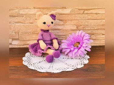Crocheted amigurumi cat with peach body and purple ruffled dress sitting on a white doily next to a purple flower