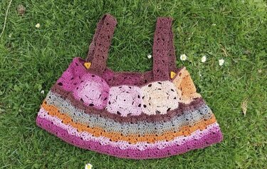 Gehäkeltes Kinderkleid in Lila-, Orange- und Brauntönen mit Streifen und runden Applikationen, auf grünem Gras liegend