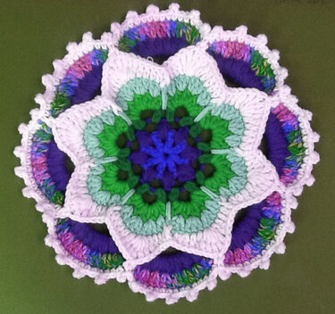 Rundes gehäkeltes Mandala in Blütenform mit Weiß, Violett, Grün, Blau und Rosa auf grünem Hintergrund.