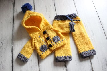 Yellow hand-knitted baby hooded jacket with blue pompom, grey patches and matching pants on white wood.
