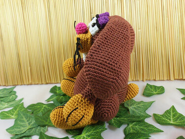 Gehäkelter Amigurumi-Hase in Braun und Gelb mit rosa Nase und lila Akzent, sitzend vor einer Holzlattenwand zwischen grünen Blättern.
