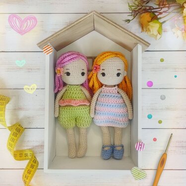 Two crocheted amigurumi dolls with braided hair and colorful outfits inside a wooden house-shaped shelf