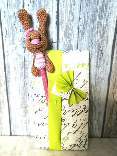 Brauner gehäkelter Amigurumi-Hase mit rosa Schleife als Stift-Topper vor verpacktem Geschenk mit grünem Band