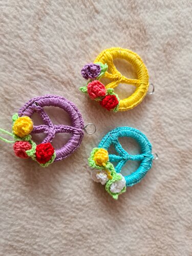 Drei gehäkelte Peace-Anhänger (gelb, türkis, lila) mit kleinen bunten Blumen auf hellem Stoff