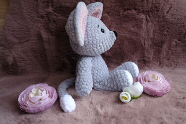 Gehäkelte graue Maus mit rosa Ohren und weißer Schwanzspitze auf braunem Plüschteppich, umgeben von rosa und weißen Stoffblüten.