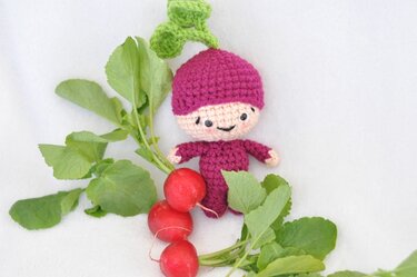 Gehäkelte Amigurumi-Figur im Radieschen-Stil mit grünem Blattgrün, neben frischen Radieschen und Blättern