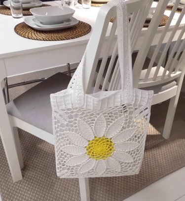 Weiße gehäkelte Shopper-Tasche mit großem Gänseblümchen hängt an einem Stuhl