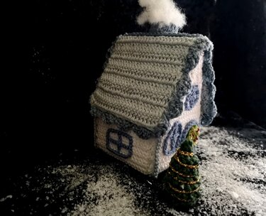 Gehäkeltes Minihaus mit hellgrauem Dach, blauen Fensterapplikationen und kleinem Tannenbaum auf Kunstschnee