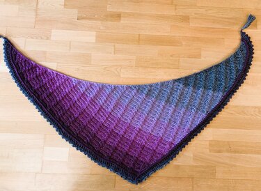 Handgehäkeltes Dreieckstuch mit Lila‑zu‑Grau‑Blau‑Farbverlauf, strukturiertes Muster, Wellenrand und Quaste, auf Holzfußboden ausgebreitet