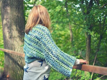 Rückenansicht einer Frau, die einen blau-grünen, offen gehäkelten Bolero trägt und an einem Holzgeländer im Wald steht.