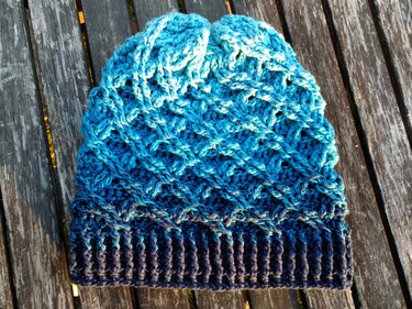 Gehäkelte Damenmütze in Blau mit Farbverlauf und Rippbündchen, liegend auf Holzdielen