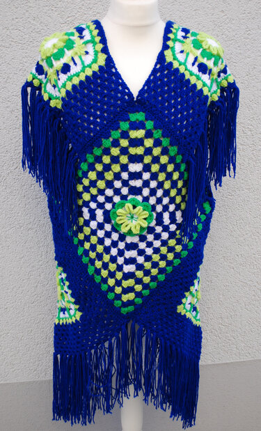 Blauer gehäkelter Poncho aus Granny-Squares mit grünen und weißen Akzenten, V‑Ausschnitt, Fransen und zentraler Häkelblume auf Schaufensterpuppe