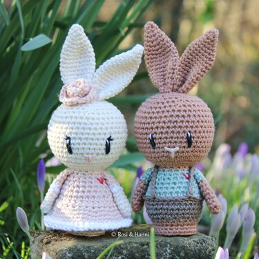 Zwei gehäkelte Amigurumi-Hasen, helles Häschen mit Kleid und braunes Häschen mit Latzhose, stehend im Frühlingsgarten