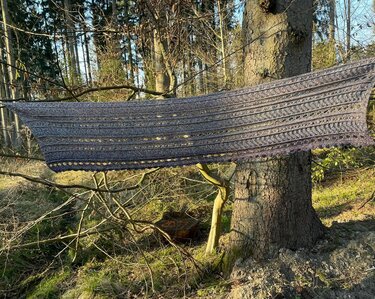 Grey-blue rectangular crochet shawl with lace and textured stitch bands hanging between tree branches.