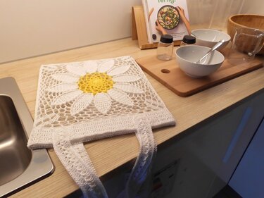 Square white crochet shopper bag with yellow daisy panel and two handles lying on a kitchen counter